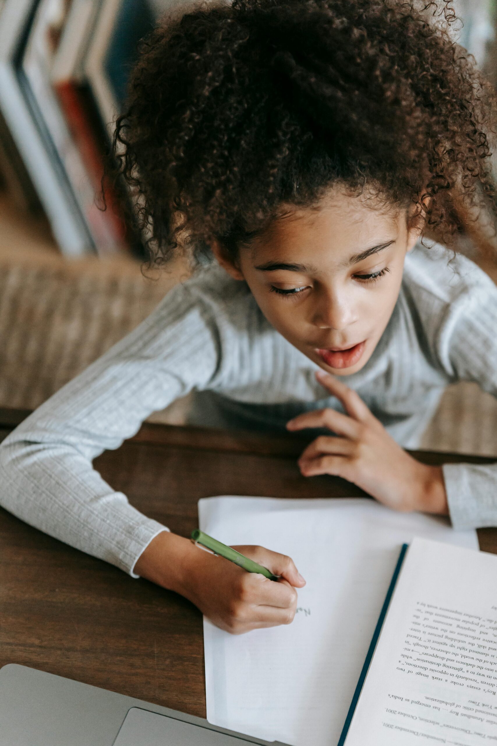 From above of attentive little black girl with Afro hair in casual clothes sitting at table with laptop and nooks while doing homework assignment